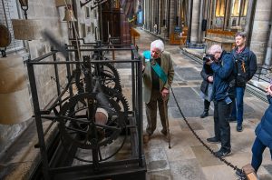 Elderly man wearing a sash gesturing at a mechanical clock, people are watching and taking photos