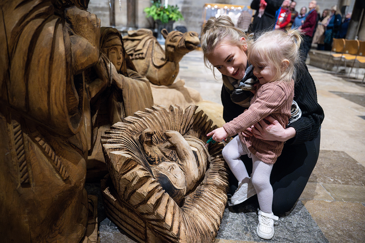 Small child and carer interact with wooden nativity scene